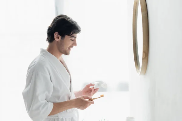 Вид сбоку человека в белом халате, стоящего с зубной пастой и зубной щеткой у зеркала — стоковое фото