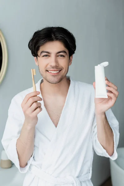 Homem feliz com escova de dentes e tubo de pasta de dentes olhando na câmera no banheiro — Fotografia de Stock