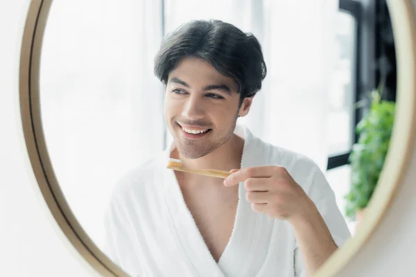 Hombre alegre en albornoz blanco de pie cerca del espejo del baño con cepillo de dientes - foto de stock