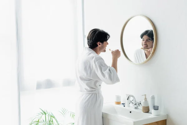 Sonriente hombre cepillarse los dientes cerca del espejo y fregadero en el baño - foto de stock