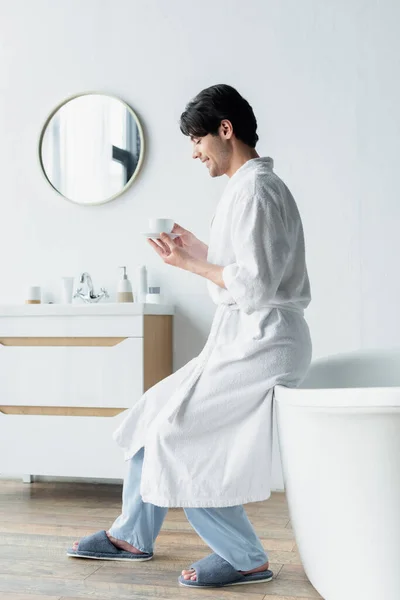 Vue latérale de l'homme heureux en peignoir assis sur la baignoire avec tasse de café — Photo de stock