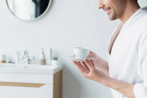 Частичный вид улыбающегося мужчины с чашкой кофе в размытой ванной комнате — стоковое фото