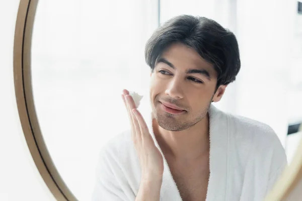 Joven hombre sonriente en albornoz aplicando espuma de afeitar cerca del espejo - foto de stock