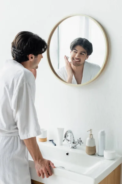 Homem sorrindo aplicando espuma de barbear perto do espelho, produtos de higiene pessoal e navalha de segurança na pia — Fotografia de Stock