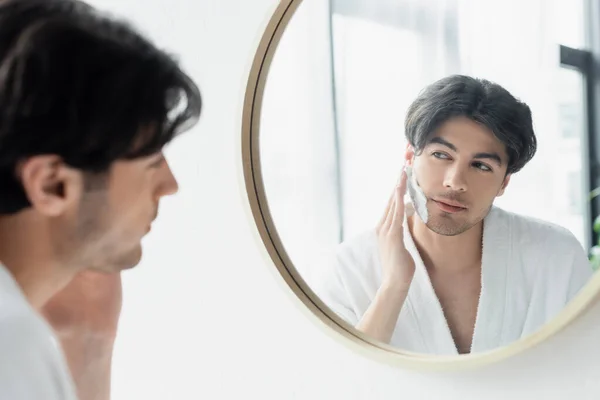 Брюнетка мужчина смотрит в зеркало ванной комнаты при нанесении пены для бритья — стоковое фото
