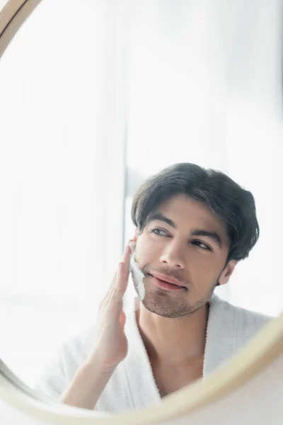 Uomo sorridente che applica schiuma da barba vicino allo specchio in bagno — Foto stock