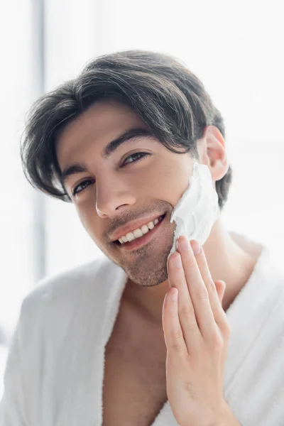 Весёлый брюнетка мужчина улыбается в камеру, нанося пену для бритья — стоковое фото