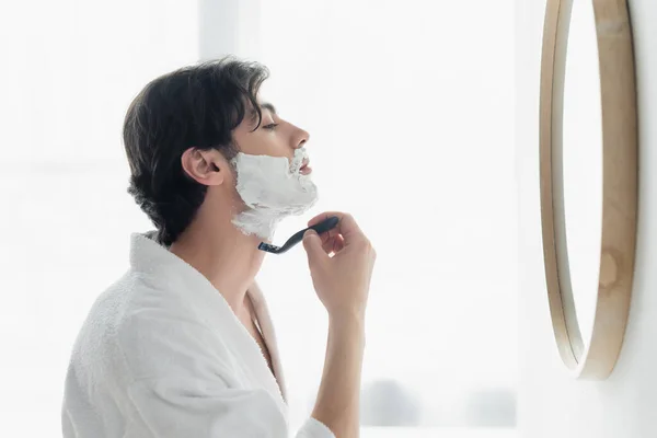 Seitenansicht eines brünetten Mannes, der sich in der Nähe eines Spiegels im Badezimmer rasiert — Stockfoto