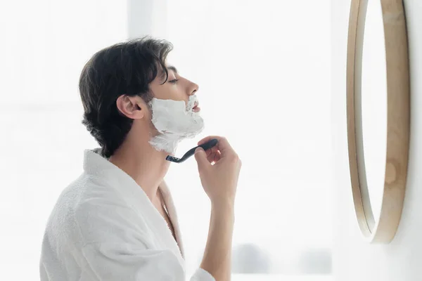 Side view of brunette man shaving near mirror in bathroom — Stock Photo
