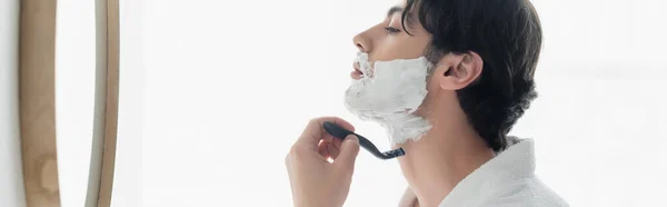 Вид збоку молода брюнетка голиться біля дзеркала у ванній кімнаті, банер — стокове фото