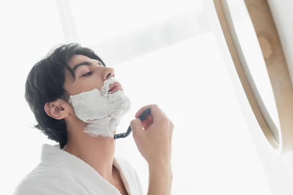 Брюнетка мужчина в белом халате бритья возле зеркала ванной — стоковое фото