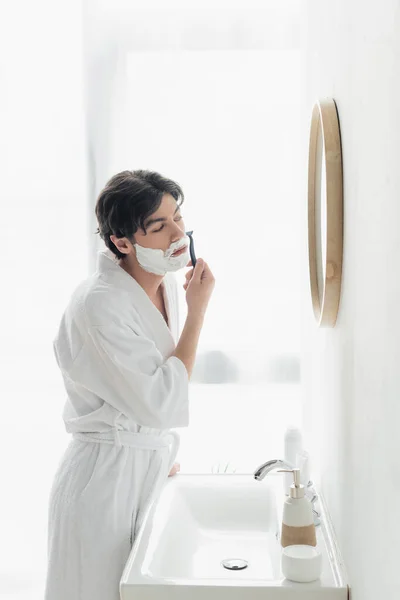 Jeune homme en peignoir blanc rasage près du miroir et lavabo dans la salle de bain — Photo de stock