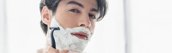 Young man shaving with safety razor and looking at camera, banner — Stock Photo