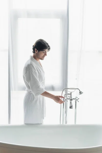 Young brunette man in white bathrobe opening faucet in bathroom — Stock Photo