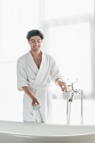 Homem sorridente em roupão de banho branco segurando cabeça de chuveiro ao abrir a torneira no banheiro — Fotografia de Stock