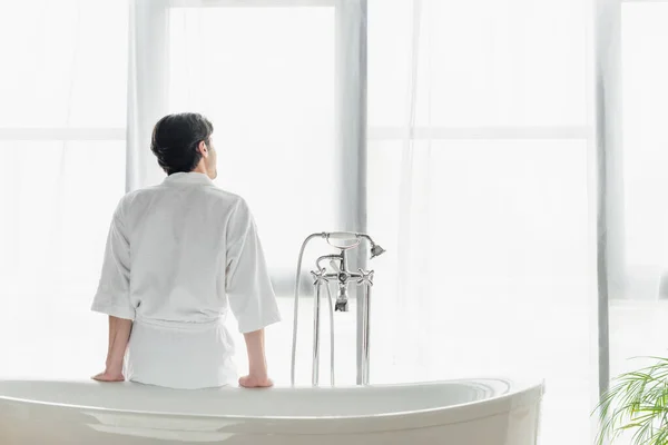 Rückansicht eines Mannes im weißen Bademantel, der auf einer Badewanne am Fenster sitzt — Stockfoto