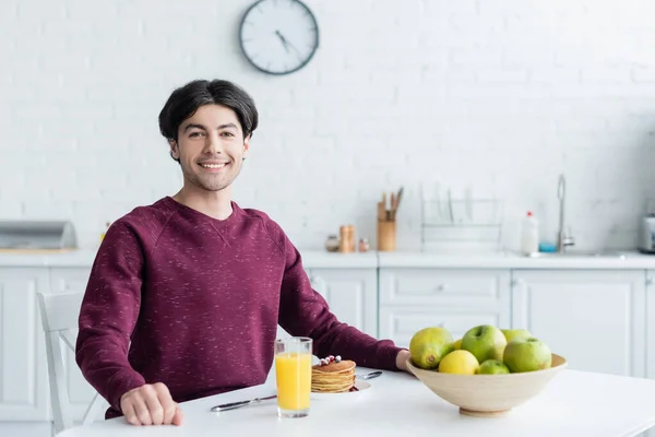 Glücklicher Mann blickt in die Kamera in der Nähe von leckerem Frühstück und frischem Obst in der Küche — Stockfoto