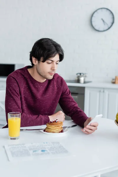 Morena hombre utilizando el teléfono móvil cerca de panqueques y zumo de naranja en la cocina - foto de stock