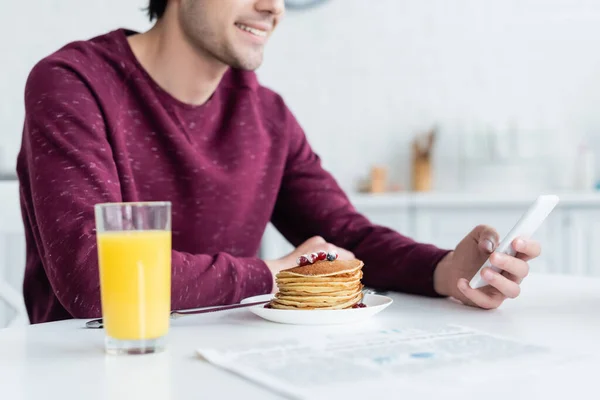 Cropped view of smiling man using smartphone near pancakes and orange juice — Stock Photo