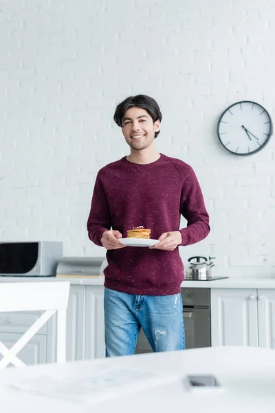 Homem morena sorrindo para a câmera enquanto segurando panquecas saborosas na cozinha — Fotografia de Stock