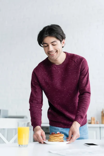 Hombre feliz sosteniendo panqueques cerca de jugo de naranja y teléfono inteligente en la mesa de la cocina - foto de stock