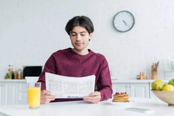 Joven leyendo el periódico matutino cerca del zumo de naranja y panqueques en la mesa - foto de stock