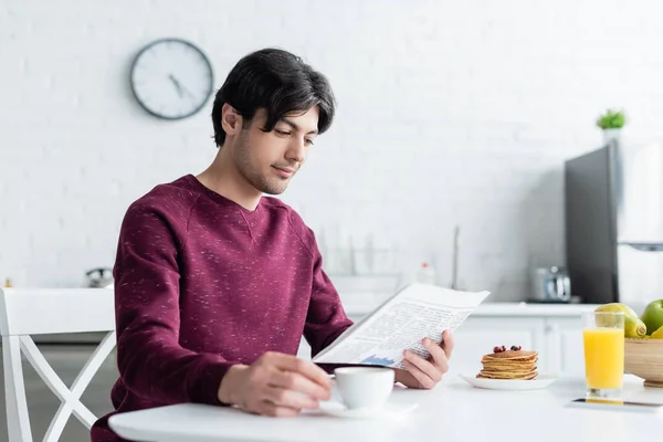 Young man reading newspaper near pancakes, orange juice and blurred coffee cup — Stock Photo