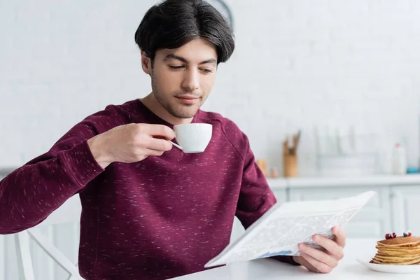 Joven bebiendo café y leyendo el periódico cerca de sabrosos panqueques en la cocina - foto de stock