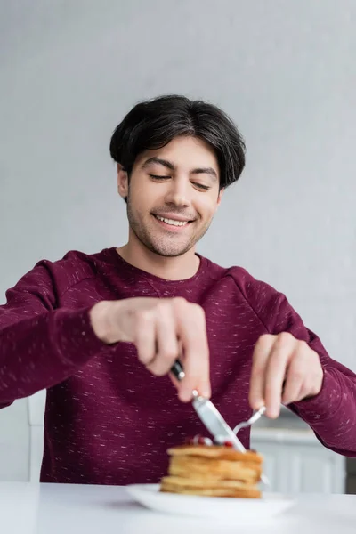 Homem alegre cortando panquecas enquanto toma café da manhã em primeiro plano desfocado — Fotografia de Stock