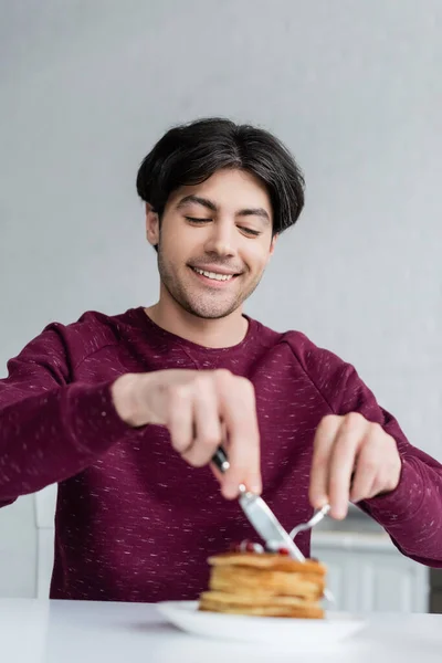 Cheerful man cutting pancakes while having breakfast on blurred foreground — Stock Photo