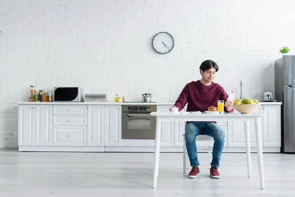 Vue pleine longueur de l'homme regardant smartphone tout en prenant le petit déjeuner dans la cuisine spacieuse — Photo de stock