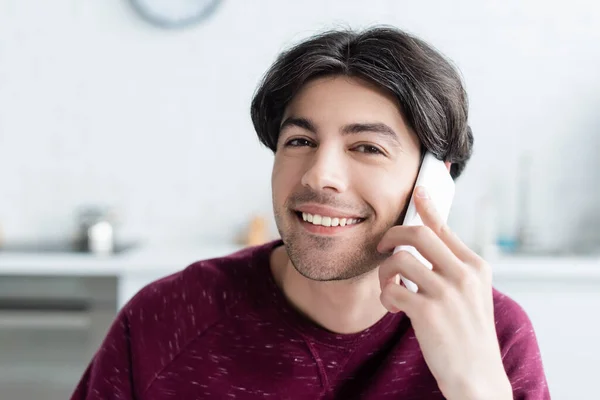Happy brunette man talking on mobile phone and looking at camera at home — Stock Photo