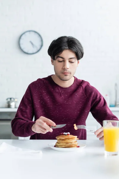 Morena hombre comiendo sabrosos panqueques cerca borrosa vaso de jugo de naranja - foto de stock