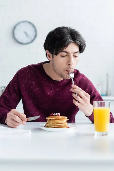 Joven morena hombre comiendo sabrosos panqueques cerca de vaso de jugo de naranja fresco - foto de stock