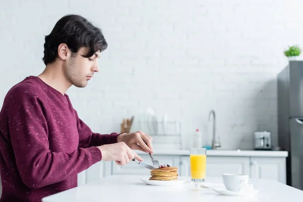 Seitenansicht eines brünetten Mannes, der Pfannkuchen in der Nähe von Orangensaft und Kaffeetasse schneidet — Stockfoto