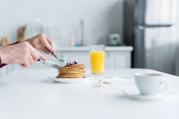 Partial view of mam cutting pancakes near orange juice and blurred coffee cup — Stock Photo