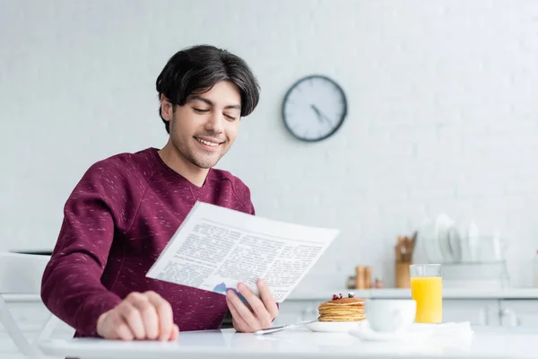 Un homme souriant lisant le journal près des crêpes et des boissons sur la table de cuisine — Photo de stock