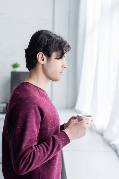 Vista laterale di giovane bruna uomo in piedi con tazza di caffè vicino alla finestra — Foto stock