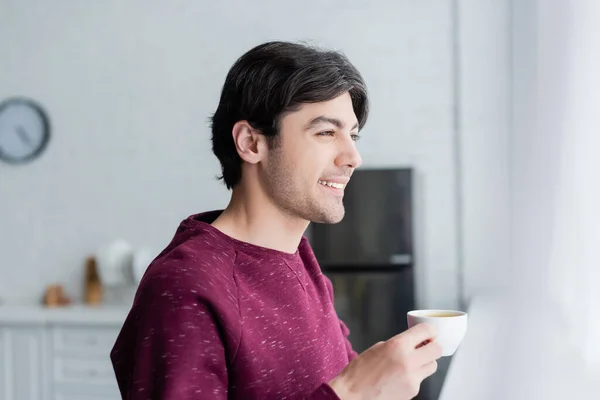 Молодой человек улыбается, держа чашку кофе в размытой кухне — стоковое фото