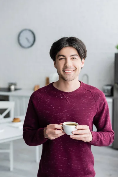 Feliz joven sosteniendo la taza de café mientras mira la cámara en la cocina - foto de stock