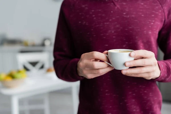 Visão parcial do homem segurando xícara de café da manhã na cozinha turva — Fotografia de Stock