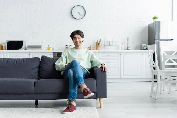 Vista completa del hombre sonriente sentado en el sofá en la cocina moderna con muebles blancos - foto de stock