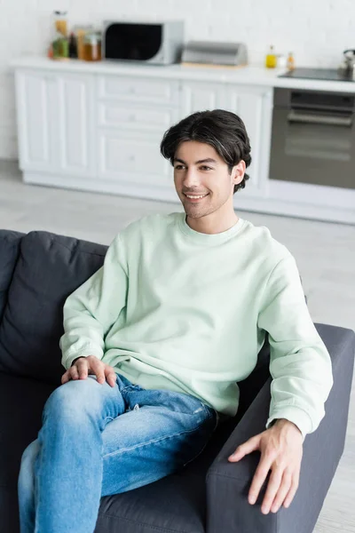 Pleased brunette man sitting on sofa in blurred kitchen — Stock Photo