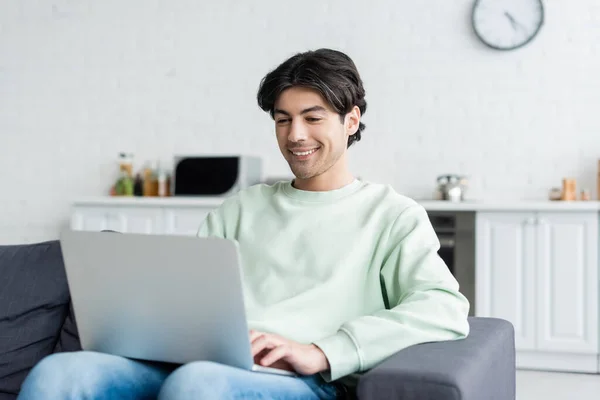 Joven feliz escribiendo en el ordenador portátil mientras está sentado en el sofá en casa - foto de stock