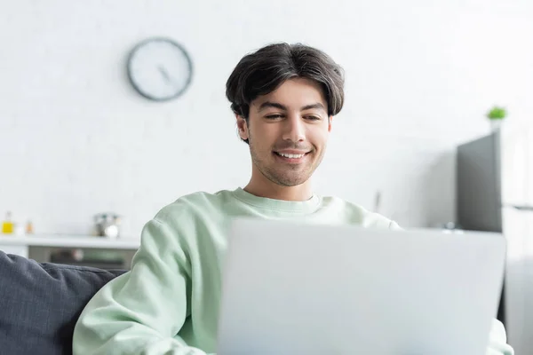 Lächelnder brünetter Mann, der zu Hause an verschwommenem Laptop arbeitet — Stockfoto