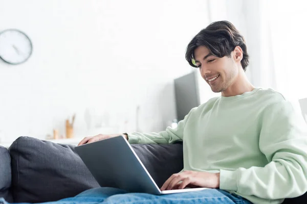 Jeune homme positif utilisant un ordinateur portable sur le canapé à la maison — Photo de stock