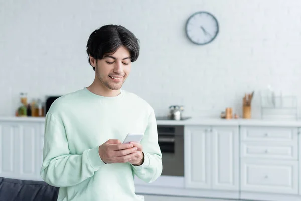 Mensagens homem alegre no telefone celular na cozinha embaçada — Fotografia de Stock