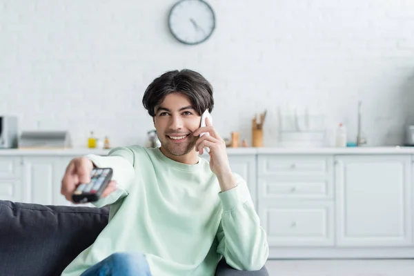 Cheerful man talking on smartphone while watching tv at home — Stock Photo