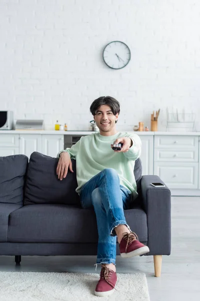 Visão comprimento total do homem feliz sentado no sofá e clicando canais de tv — Fotografia de Stock