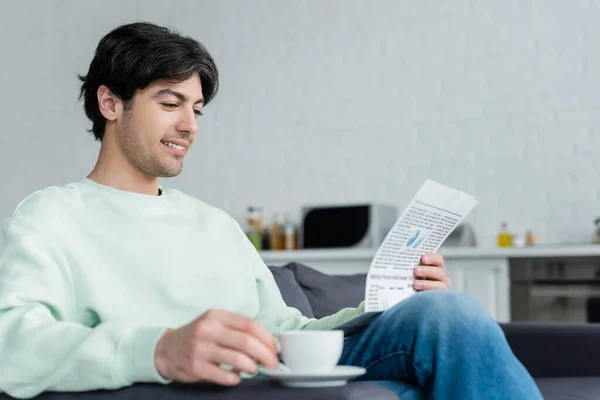 Un homme souriant lisant un journal près d'une tasse de café floue sur un canapé — Photo de stock